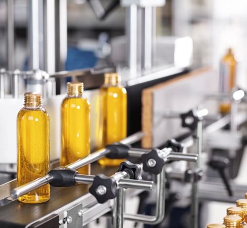 Botellas de plástico llenas de líquido ámbar avanzando por una cinta transportadora industrial.
Se ven guías y soportes metálicos que alinean los envases durante el proceso.
Al fondo aparece maquinaria de envasado o etiquetado desenfocada en una línea de producción.
