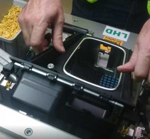 Manos de un operario manipulando una máquina o estación de trabajo industrial.
Se observa un módulo con carcasa negra y un teclado o panel de botones integrado.
La escena muestra una intervención de ajuste o mantenimiento sobre el equipo en planta.
