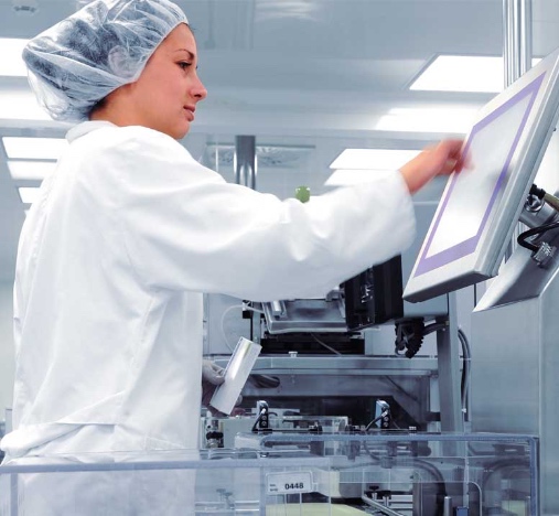 Operaria en entorno industrial con bata blanca y redecilla, usando una pantalla táctil de control.
La persona interactúa con un panel HMI montado en una máquina de producción automatizada.
Al fondo se ven equipos y protecciones transparentes en una planta limpia e iluminada.
Imagen asociada al control y supervisión de procesos en fábrica.
