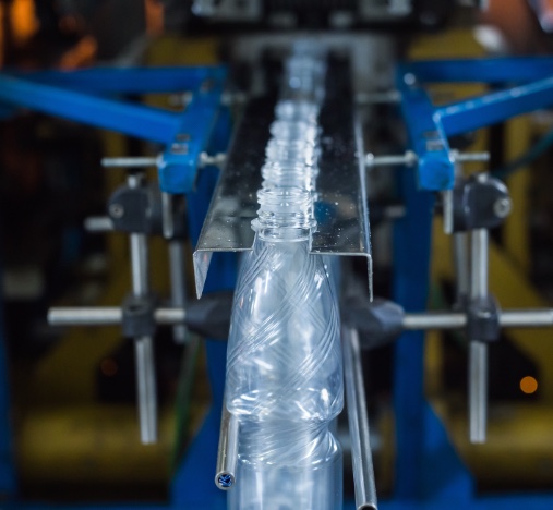 Botellas de plástico transparentes avanzando por una guía metálica en una línea de producción.
Se ven brazos y soportes azules de la maquinaria que alinean los envases durante el proceso.
Imagen de un entorno industrial de fabricación o envasado.
