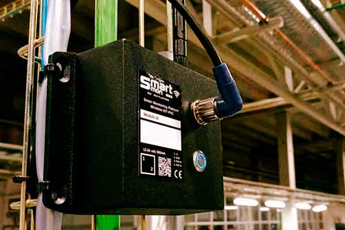 Módulo de SmartMon montada en una estructura metálica dentro de una fábrica.
                                             Se ve una antena conectada al lateral y una etiqueta con el logo, código QR y datos del equipo.
                                             Al fondo aparecen tuberías y vigas del techo, con iluminación industrial desenfocada.
                                             Imagen que sugiere un dispositivo de monitorización/IoT instalado en planta.
                                             