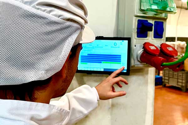 Operario con ropa y gorro de trabajo usando una tablet industrial en planta.
                                             En la pantalla se muestra SmartMon en el módulo de producción, con gráficos y barras de estado.
                                             La tablet está montada junto a conectores y mandos industriales en una pared de la fábrica.
                                             Escena de control y seguimiento de la producción en tiempo real.
                                             